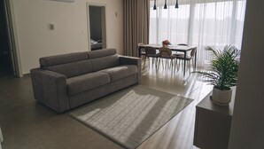 Un salon moderne avec un canapé gris, une table à manger avec des chaises, une plante en pot et une grande fenêtre avec des rideaux.