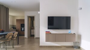 Een moderne woonkamer met een flatscreen televisie aan de muur, een eethoek met een tafel en stoelen, en een keuken op de achtergrond.