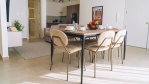 Een moderne eetruimte met een tafel voor vier personen, omringd door stoelen met gouden poten. Op de tafel staat een fruitschaal, en er is een keuken zichtbaar op de achtergrond.