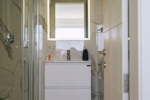 Ein Badezimmer mit Duschkabine aus Glas, einem weißen Waschbecken mit Spiegel darüber und Wänden mit Marmoroptik.