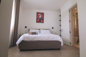 Ein modernes Schlafzimmer mit einem großen Bett, einem Nachttisch, einem kleinen Tisch daneben, einem gerahmten Bild an der Wand und einem durch eine offene Tür einsehbaren Badezimmer.