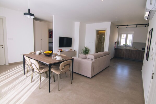 Eine moderne Küche mit Flachbildfernseher, ein Essbereich mit Holztisch und -stühlen sowie eine Wohnarea mit Sofa und Pflanze.