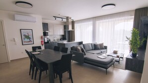 A modern living room with a dining area, a kitchen in the background, and a large window with sheer curtains. - The Valley Collection - Suite A10 (Ix-Xagħra)