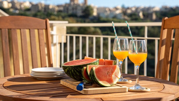 A wooden table with a watermelon and two glasses of orange juice on a balcony with a city view. - The Valley Collection - Suite B11 (Ix-Xagħra)