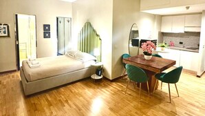 Ein modernes Hotelzimmer mit Bett, kleinem Tisch mit Lampe, Essbereich mit Holztisch und Stühlen sowie einer Küchenzeile.