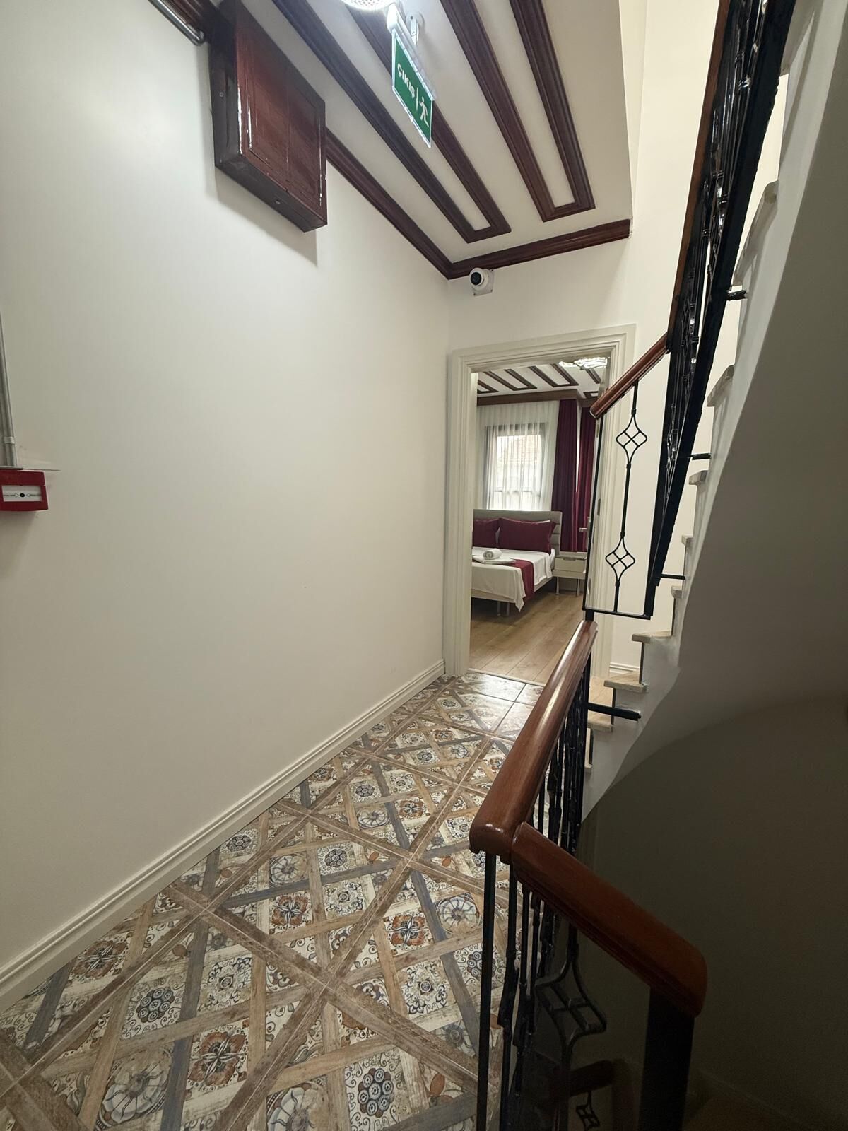 Un passadís amb rajoles de terra estampades, una escala amb barana de fusta, i una habitació amb un llit i cortines al fons.