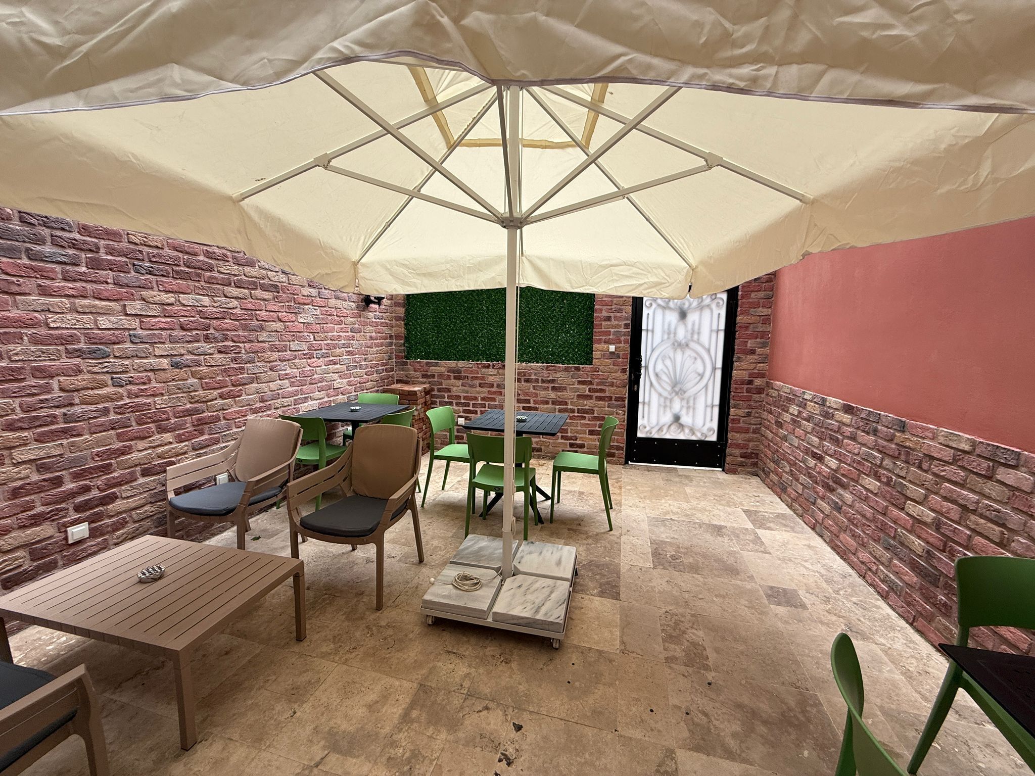 An outdoor patio with brick walls, green chairs, wooden tables, and a large umbrella.