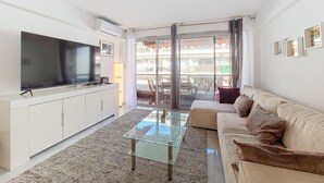 A modern living room with a flat-screen TV, a glass coffee table, a beige sofa, and a view through a glass door to an outdoor balcony. - N&J - CLEA - Terrace - Central (Niza)