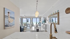Et moderne køkken med en hvid marmor bordplade, en guldfarvet hane og udsigt til et spiseområde med et hvidt bord og stole.