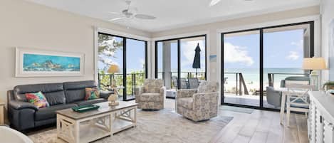 A spacious living room with a large window overlooking the ocean, a ceiling fan, and a sofa with colorful pillows.