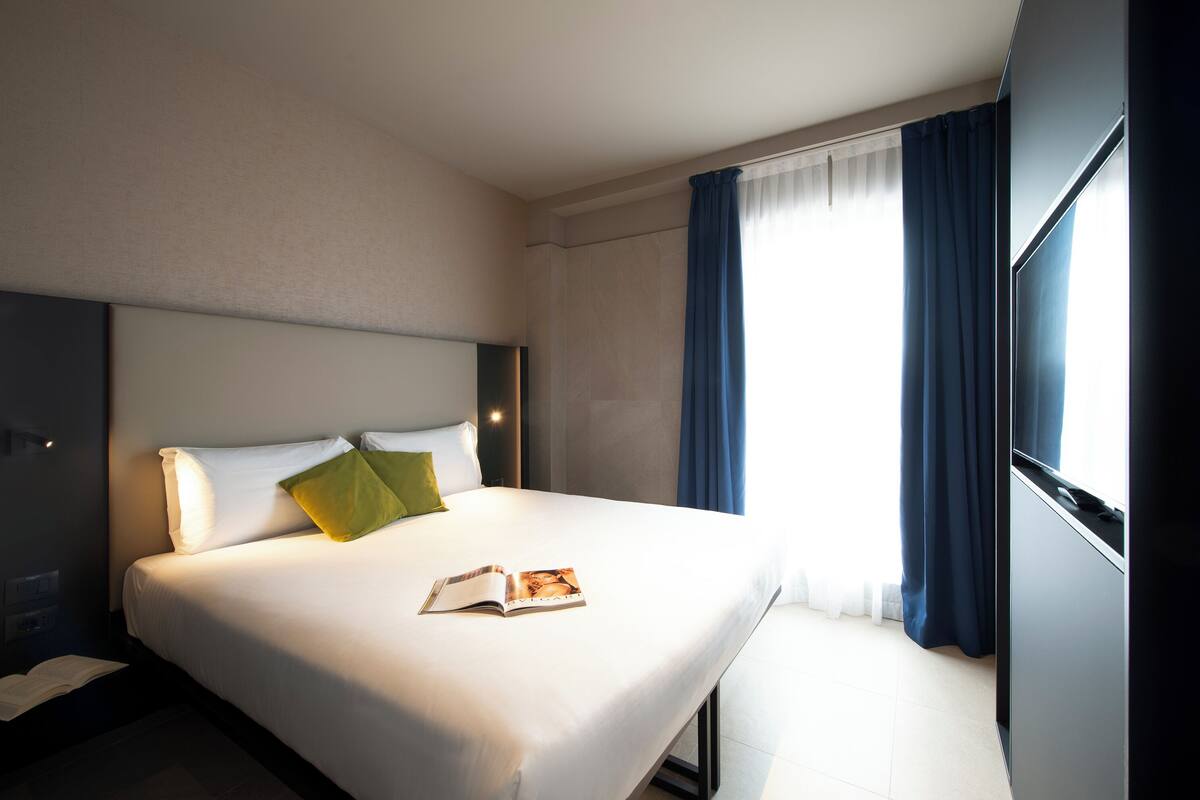 Una habitación de hotel con una cama bien hecha, una mesita de noche y una ventana con cortinas.
