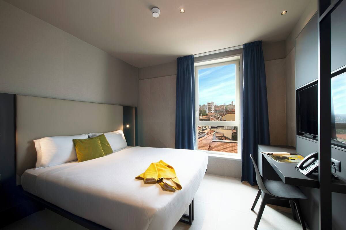 Habitación de hotel con cama, escritorio con teléfono y ventana con cortinas.