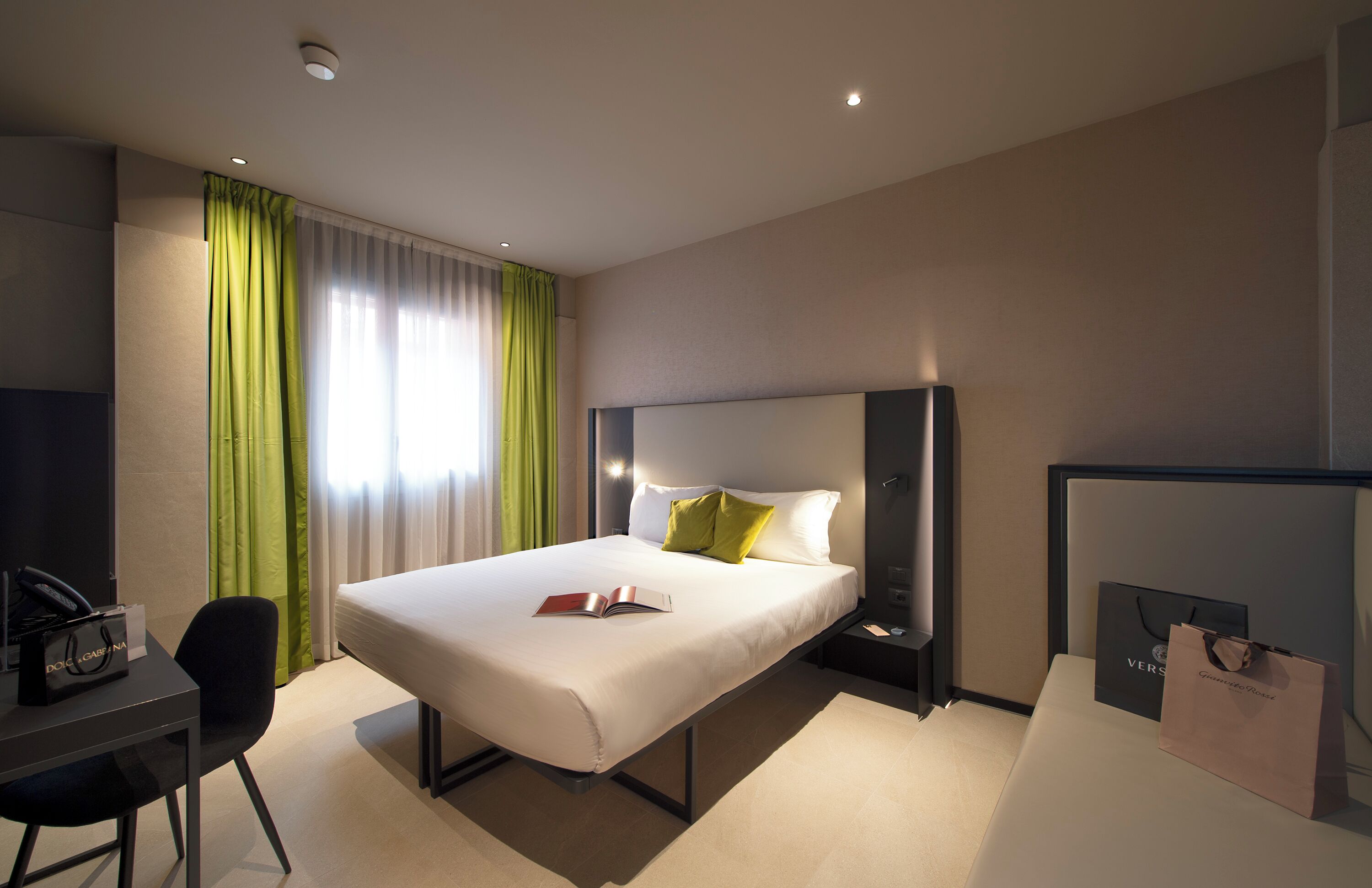A hotel room with a bed, a desk, a chair, a nightstand, a lamp, and a window with curtains.
