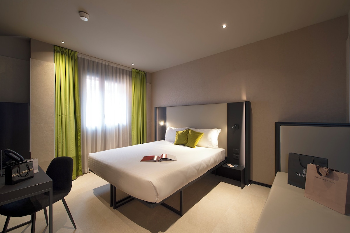 A hotel room with a bed, a desk, a chair, a nightstand, a lamp, and a window with curtains.