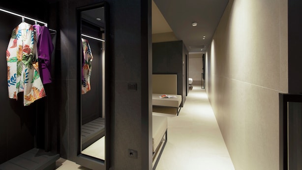Un pasillo moderno de hotel con un espejo que refleja el interior, una cama y un baño a lo lejos.