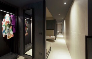 Um corredor moderno de hotel com um espelho refletindo o interior, uma cama e um banheiro visíveis ao fundo.