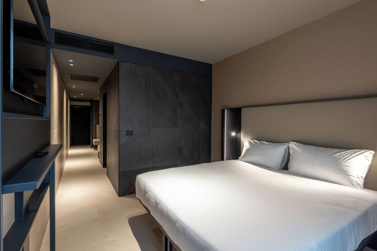 Una habitación de hotel moderna con una cama grande, una mesita de noche y televisión integrada.