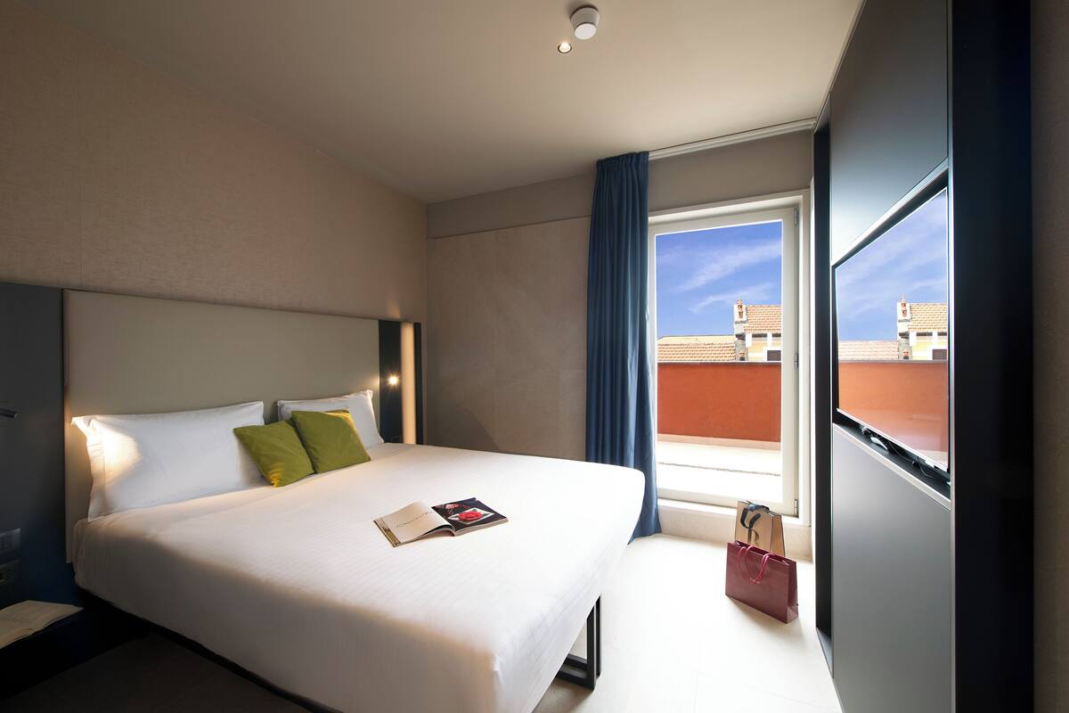 Habitación de hotel con una cama grande, una ventana con vistas a edificios y una mesita de noche con una lámpara.