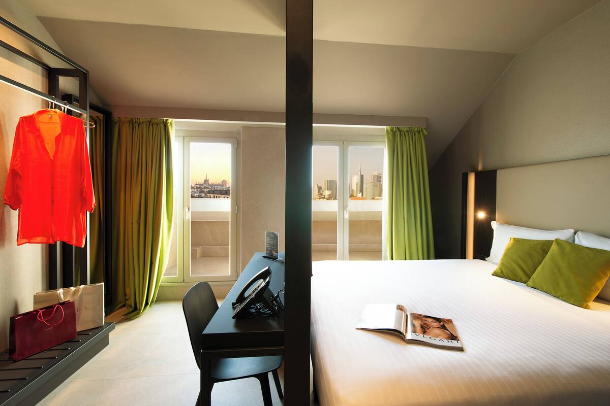 Habitación de hotel con cama, escritorio, silla, una prenda roja colgada y vista a un paisaje urbano a través de amplios ventanales.