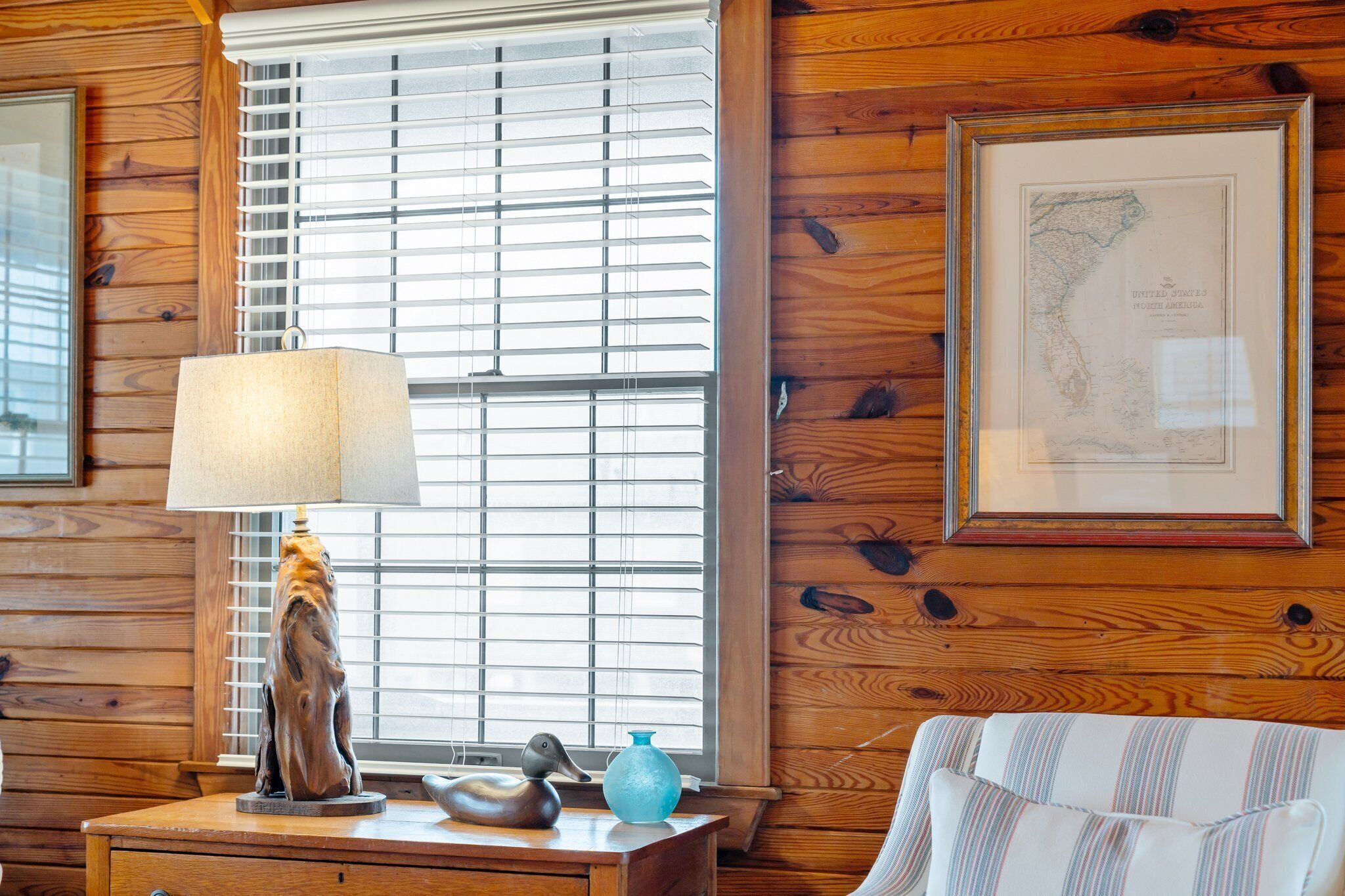 Een houten interieur met een raam, een lamp, een ingelijste kaart en een gestreept kussen.