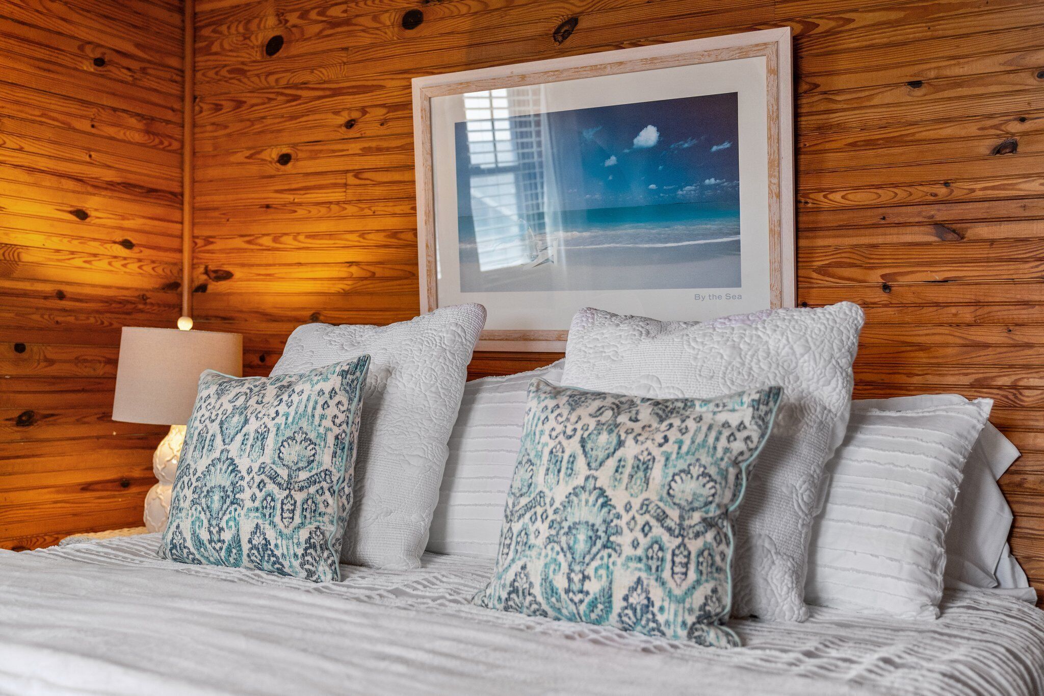 Een kamer met houten lambrisering, een bed met gekleurde kussens, een ingelijst strandbeeld en een bedlampje.