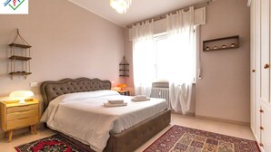 A bedroom with a large bed, bedside tables, a radiator, a window with curtains, and a patterned rug. - A Casa Di Giò by TuscanyWeGo (Livorno)