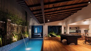 Un espace piscine intérieur moderne, doté d’une terrasse en bois, d’une table de billard et d’un mur en pierre agrémenté de plantes.