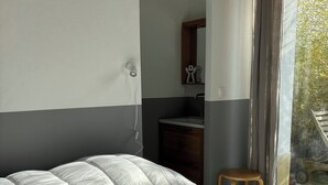 A bedroom with a large bed, a small stool, a bathroom with a mirror and sink, and a view of the outdoors through a window. - Vacation home, 300M BEACH (CABOURG town center)-6 people, 3 bedrooms (Cabourg)