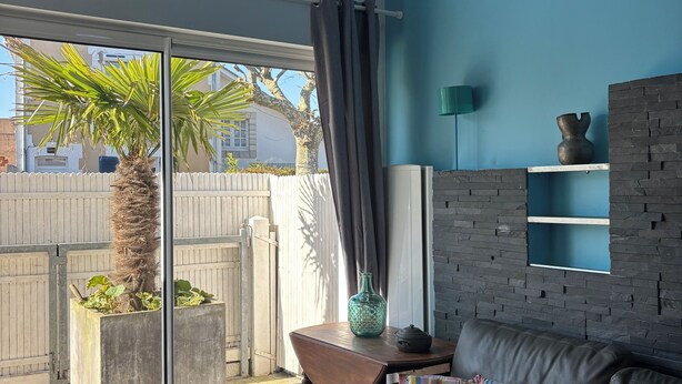 A modern living room with a large sliding glass door leading to a patio, a dark leather sofa with colorful pillows, a green vase, and a brick accent wall. - Vacation home, 300M BEACH (CABOURG town center)-6 people, 3 bedrooms (Cabourg)