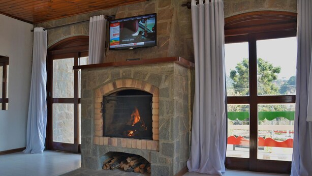 A stone fireplace with a TV mounted above it, large windows with curtains, and a view of the outdoors. - Villaggio di Roma Suítes  (Campos do Jordao)