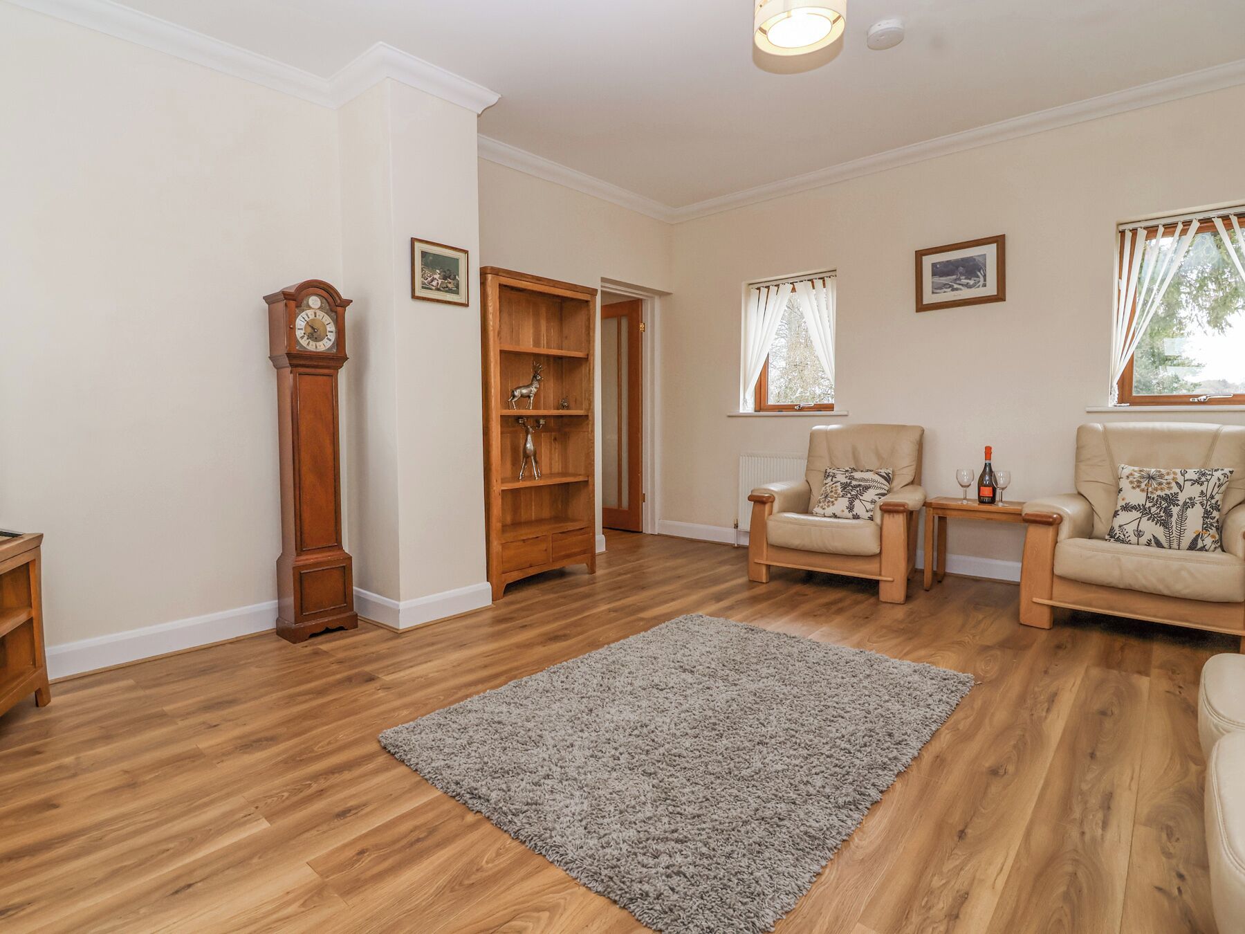 A living room with wooden flooring, a grey rug, two armchairs, a side table with wine glasses, a clock, and a bookshelf.
