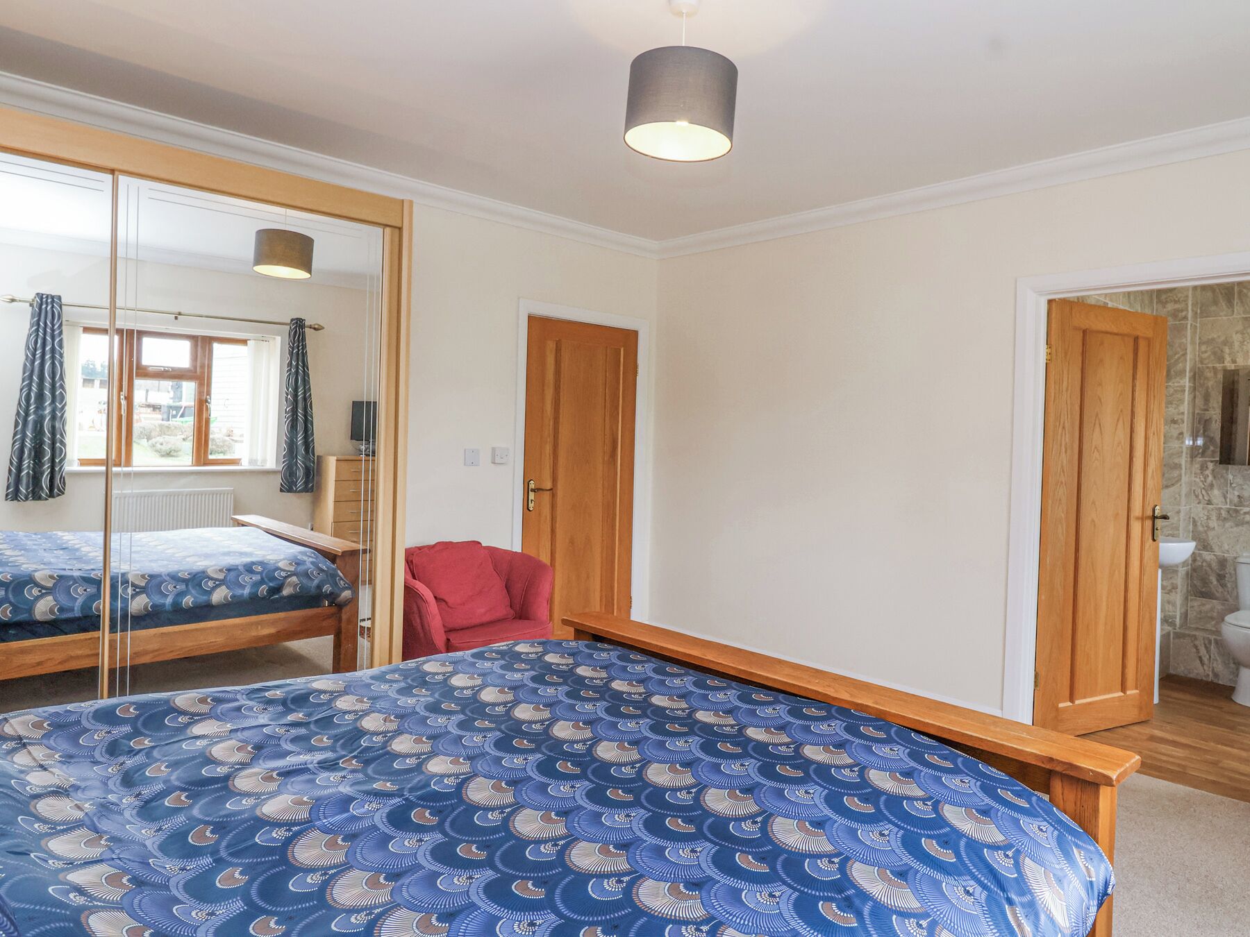 A bedroom with a bed, a mirror, a red chair, and a bathroom visible through an open door.
