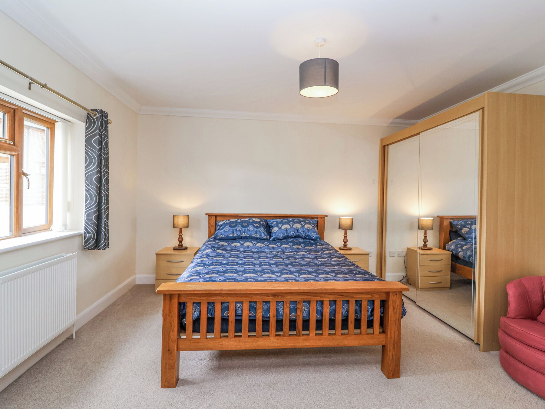 Ein Schlafzimmer mit einem hölzernen Bett, zwei Nachttischen mit Lampen, einem Fenster mit Vorhängen, einem Heizkörper und einem Kleiderschrank.