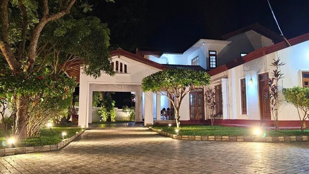Un edificio bien iluminado con entrada pavimentada, rodeado de vegetación y árboles.