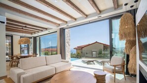 Un salon moderne avec un canapé, une table ronde en bois et des chaises, de grandes fenêtres offrant une vue sur un immeuble et une piscine.