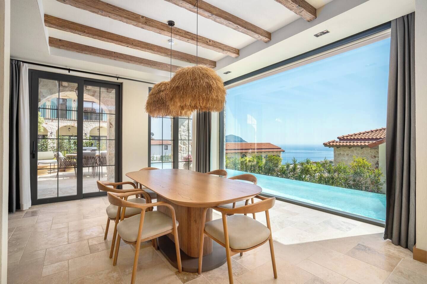 A dining area with a wooden table and chairs, large windows with a view of the sea, and a hanging light fixture.