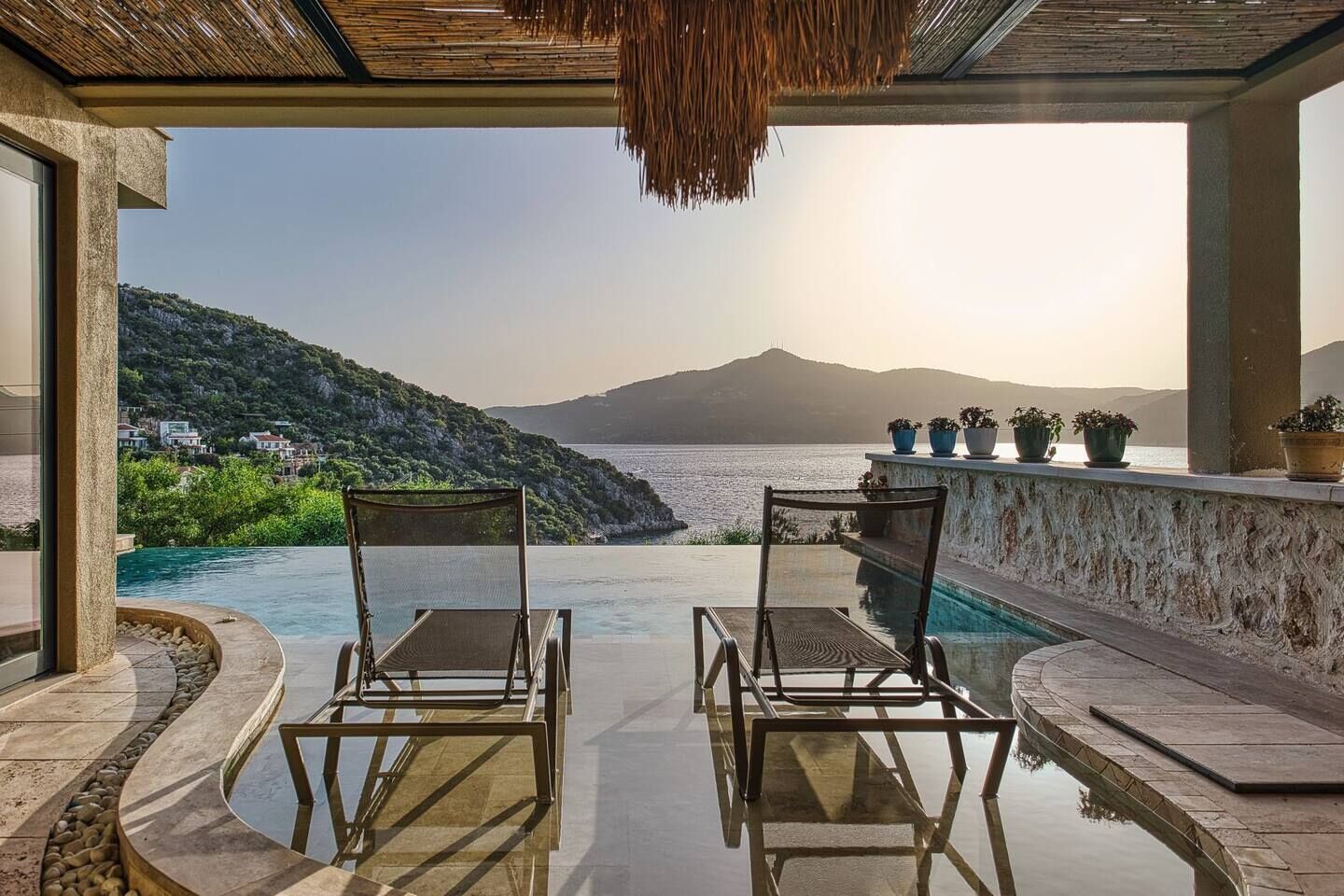 A poolside area with two lounge chairs, a view of the sea, and potted plants on a balcony.