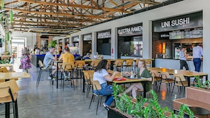 A modern dining area with wooden beams on the ceiling, multiple seating arrangements, and various restaurant signs including "UNI SUSHI". - Pool with Sea View | WI-FI 200MB | Foodhall #TT3 (Florianópolis)