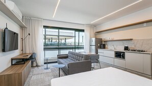 A modern hotel room with a sofa, a bed, a small table, a TV, and a kitchen area with a refrigerator, stove, and microwave. - Pool with Sea View | WI-FI 200MB | Foodhall #TT3 (Florianópolis)