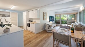 Une cuisine moderne avec des meubles blancs, un coin repas avec une table en bois dressée pour six personnes, et un coin salon confortable avec un canapé et une table basse.