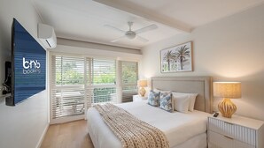 Une chambre à coucher comprenant un lit, des lampes de chevet, un ventilateur de plafond, un téléviseur et une fenêtre avec des stores.