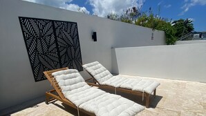 Two wooden lounge chairs with white cushions on a tiled patio against a white wall with a black leaf-patterned panel. - Sea Breeze Tunnel Hideaway 7 (Quebradillas)