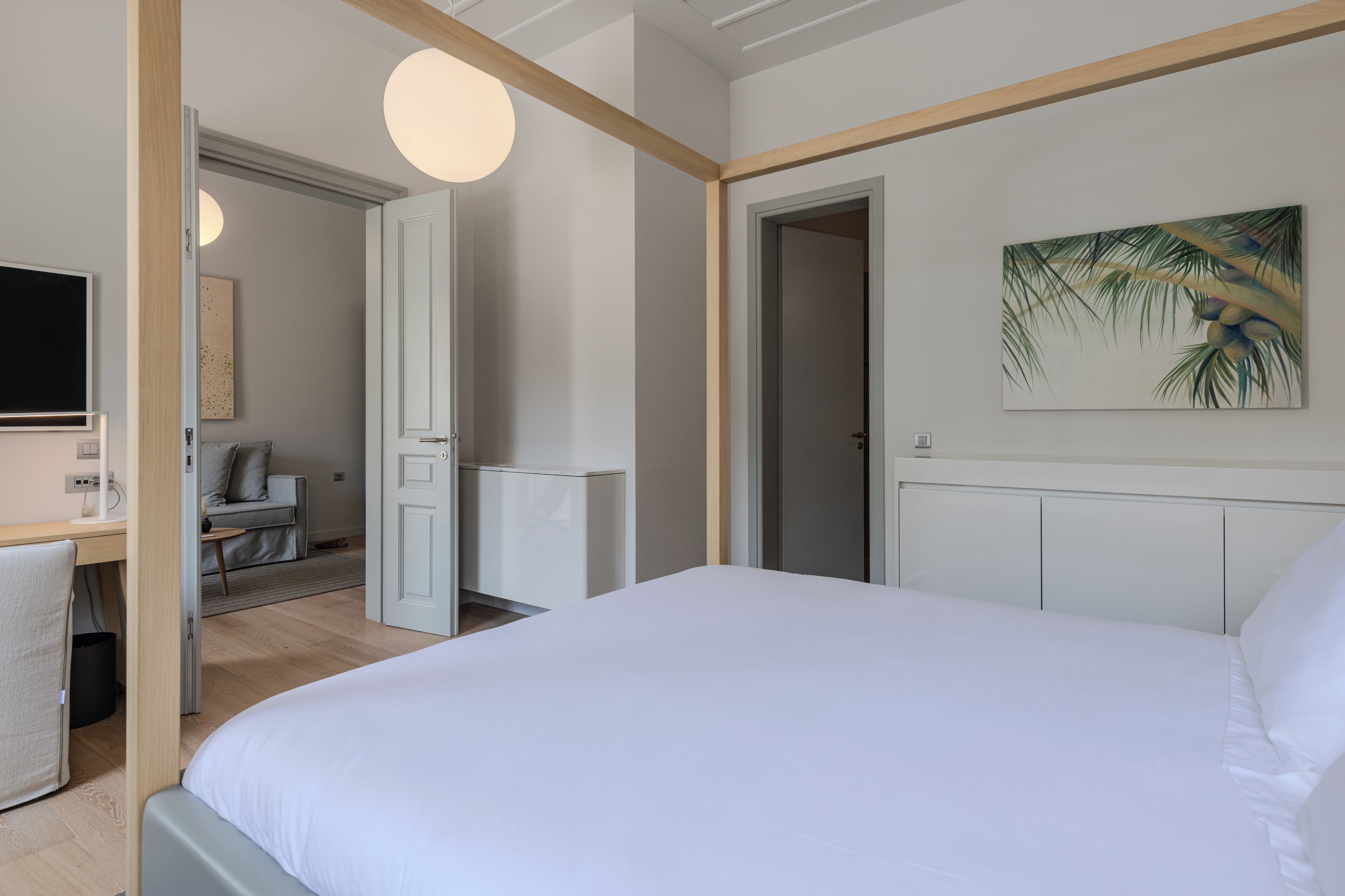 A modern hotel room with a large bed, a desk with a chair, a television, and a painting of palm leaves on the wall.