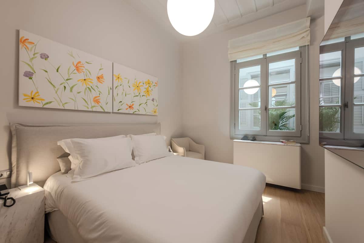 A bedroom with a large bed, a nightstand, a chair, and a window with a view of greenery.