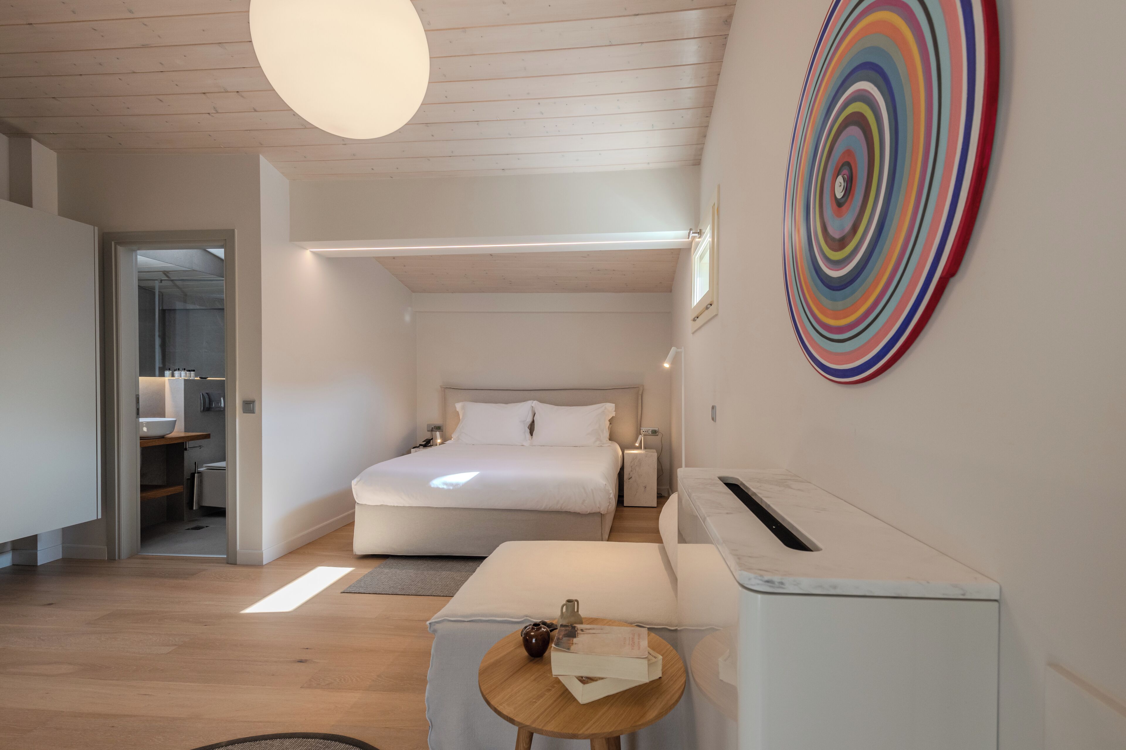 Un dormitorio moderno con cama, mesitas de noche, una pequeña mesa de madera y una obra de arte circular y colorida en la pared.