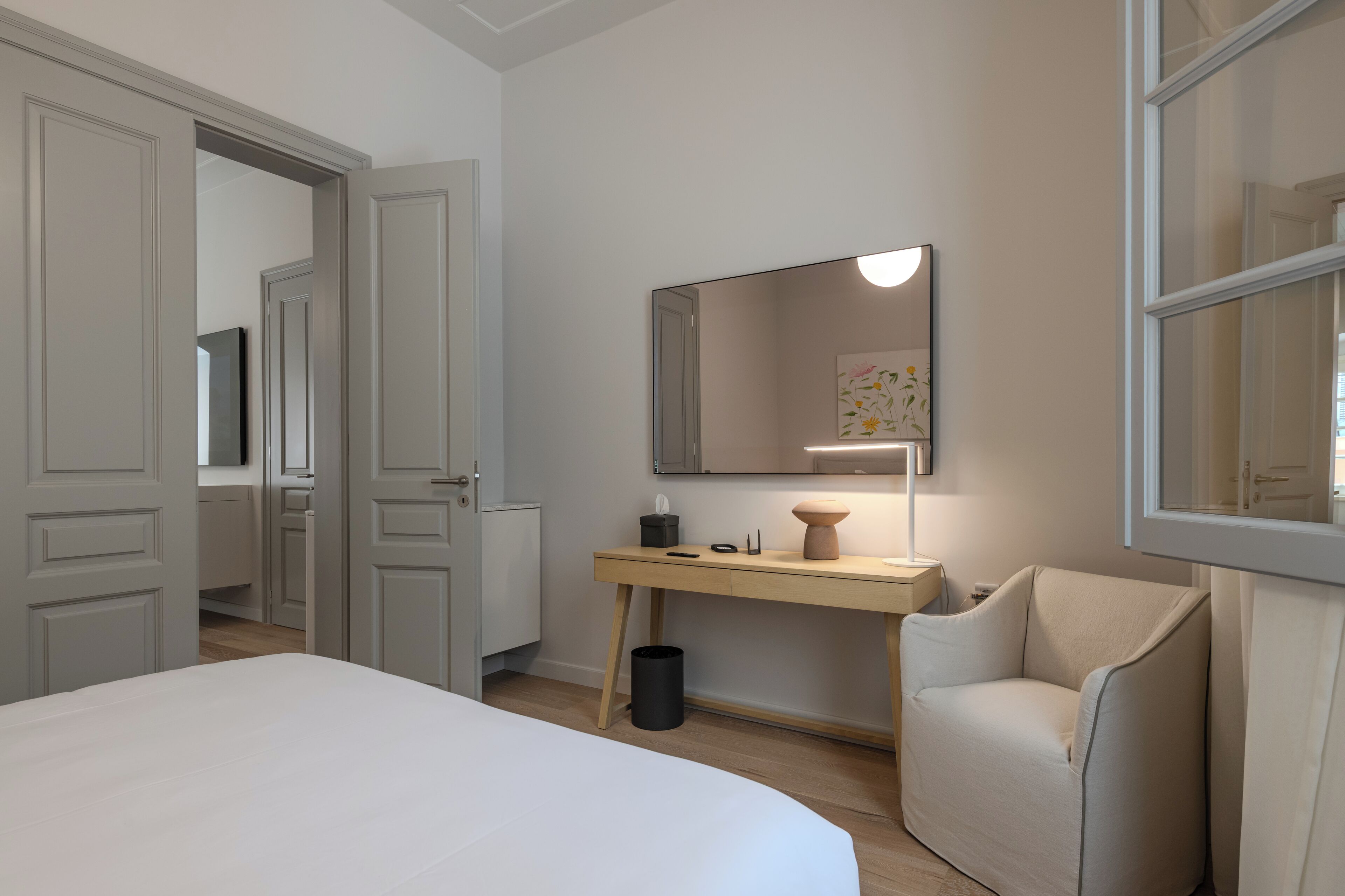 A bedroom with a bed, a desk, a chair, a mirror, and a trash can.