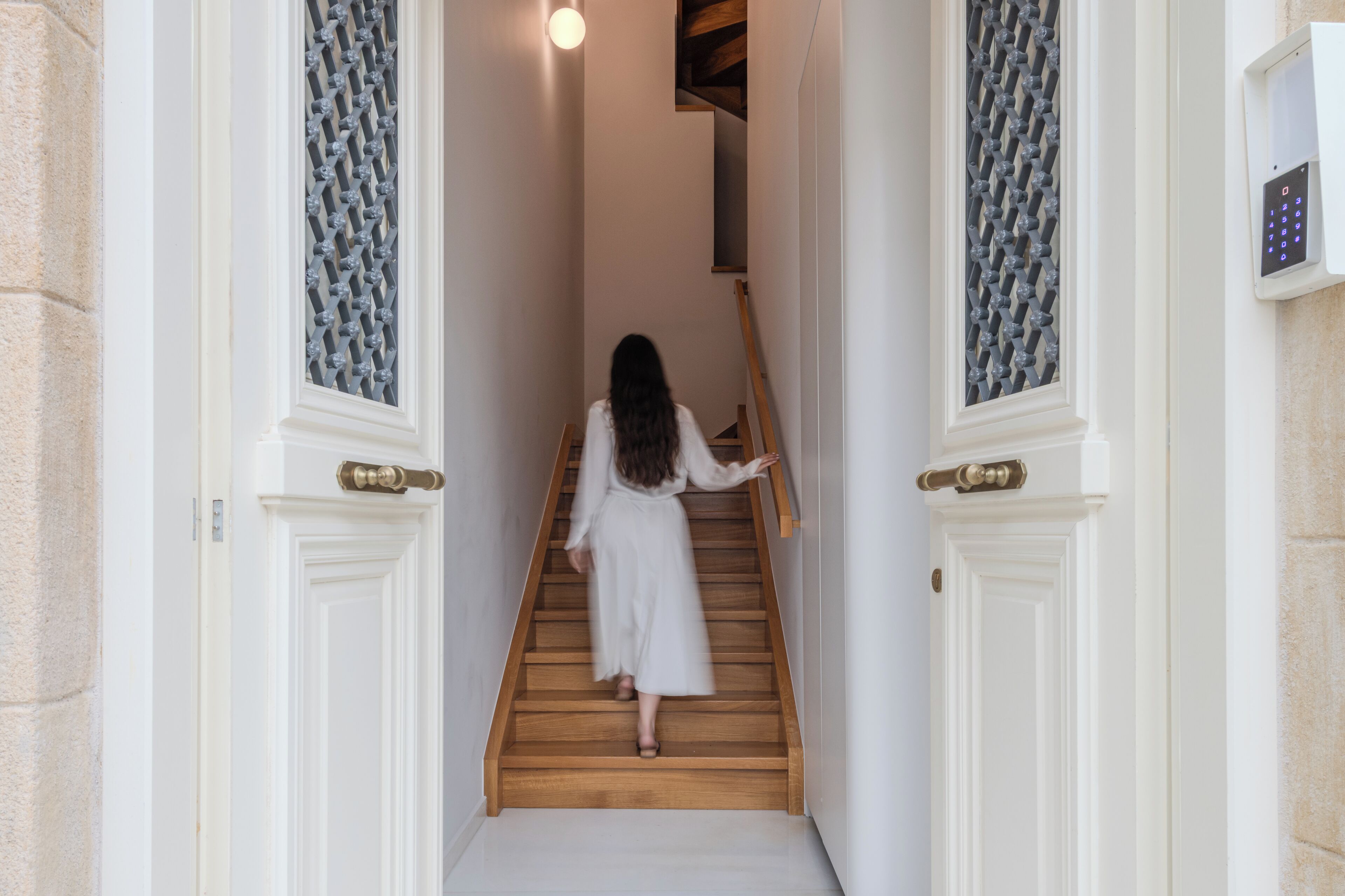 Een vrouw daalt af een houten trap in een gang met witte deuren en een beveiligingspaneel aan de muur.
