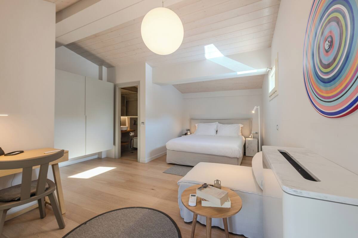 A modern hotel room with a bed, a desk, and a sofa. There is a colorful artwork on the wall and a ceiling light.