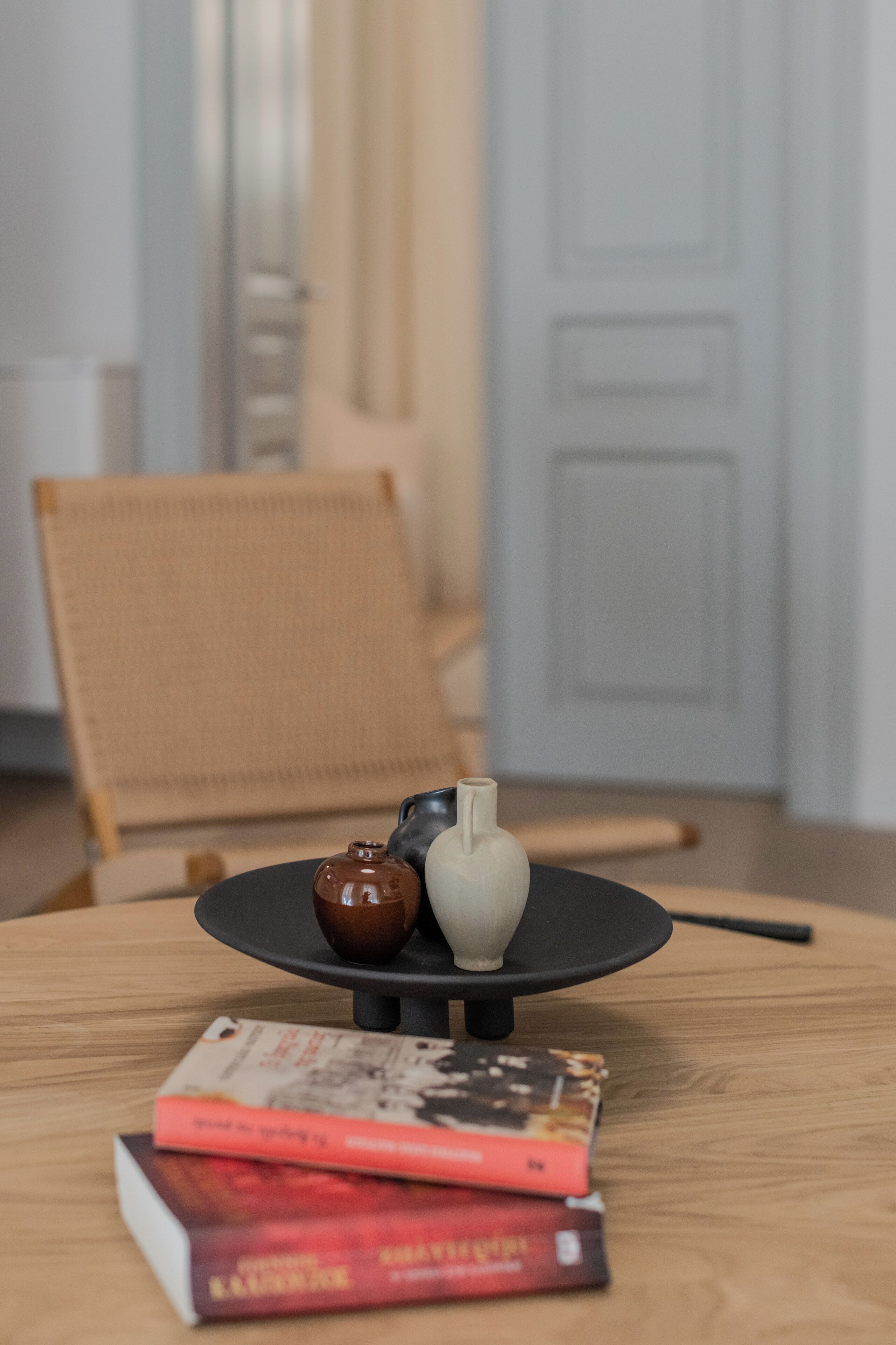 A round wooden table with a black base, a small brown vase, a white vase, and a stack of books.