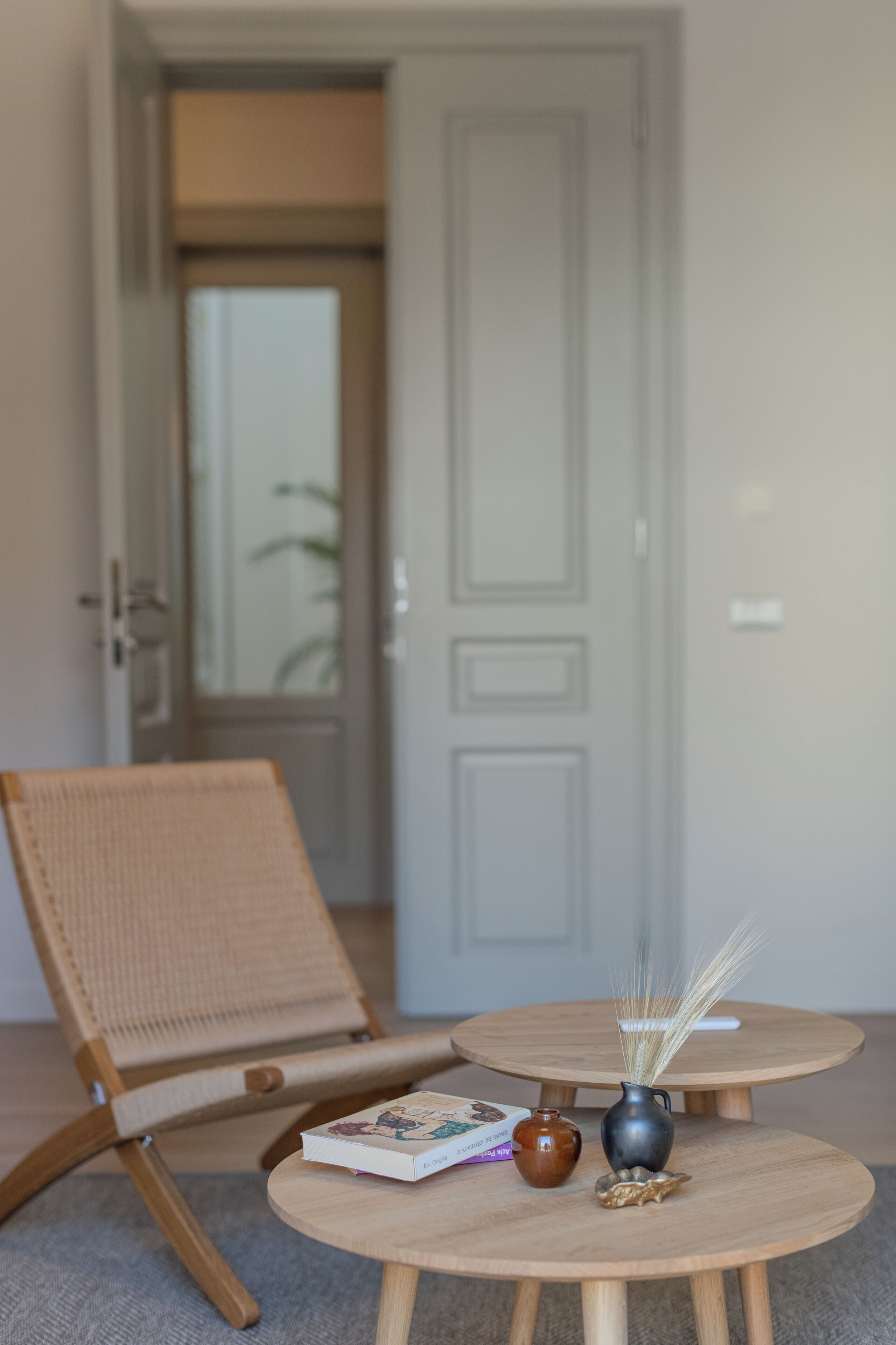 A room with a wicker chair, a small round wooden table with a book and a vase, and a door in the background.